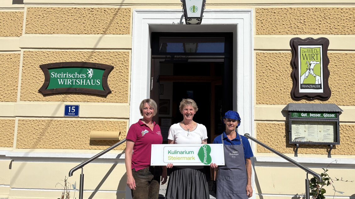 Drei Personen stehen vor einem Gasthaus mit dem Schild „Steirisches Wirtshaus“. Sie halten ein Schild mit der Aufschrift „Kulinarikum“. | © TV Hochsteiermark