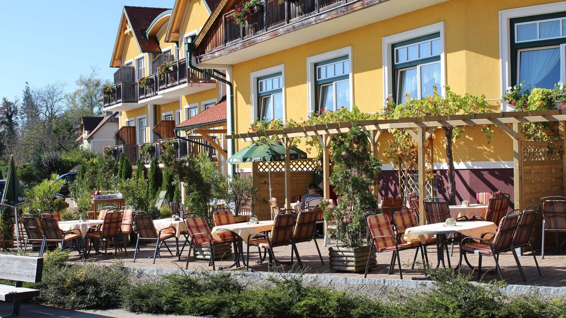 Ein gemütlicher Außenbereich mit vielen Tischen und Stühlen vor einem gelben Gebäude. Über den Tischen wachsen Pflanzen und es gibt Sonnenschirme zum Schutz vor der Sonne. | © Großschedl