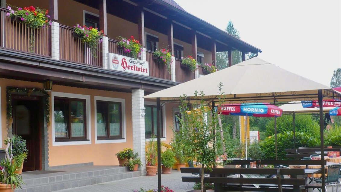 Eine gemütliche Gasthausfassade mit blühenden Blumenkästen. Draußen steht ein überdachter Sitzbereich mit Holztischen und Stühlen. | © Herlwirt