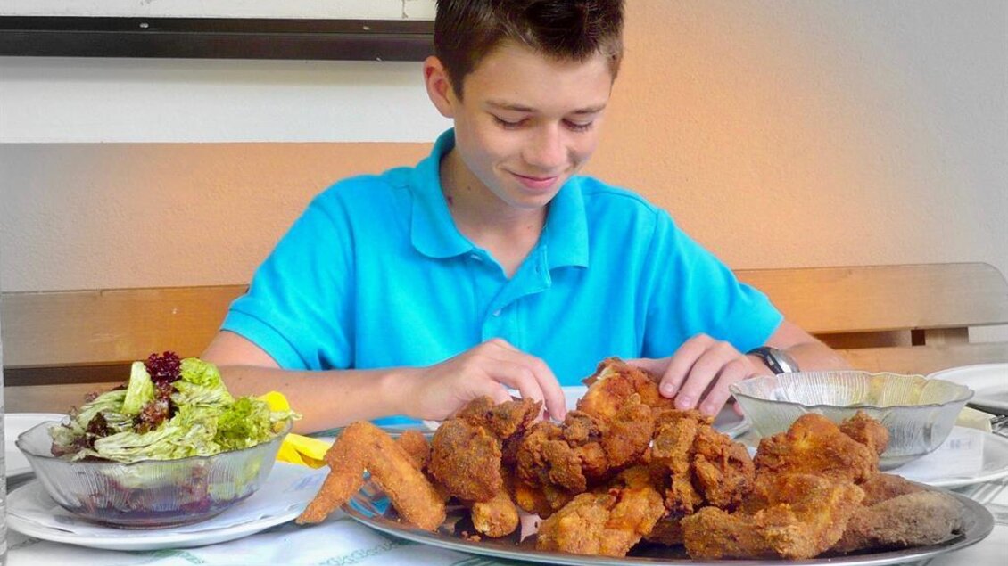 Ein Junge sitzt an einem Tisch mit einer großen Portion frittiertem Hähnchen und einem Salat. Er sieht erfreut aus und bereitet sich darauf vor, zu essen. | © Herlwirt
