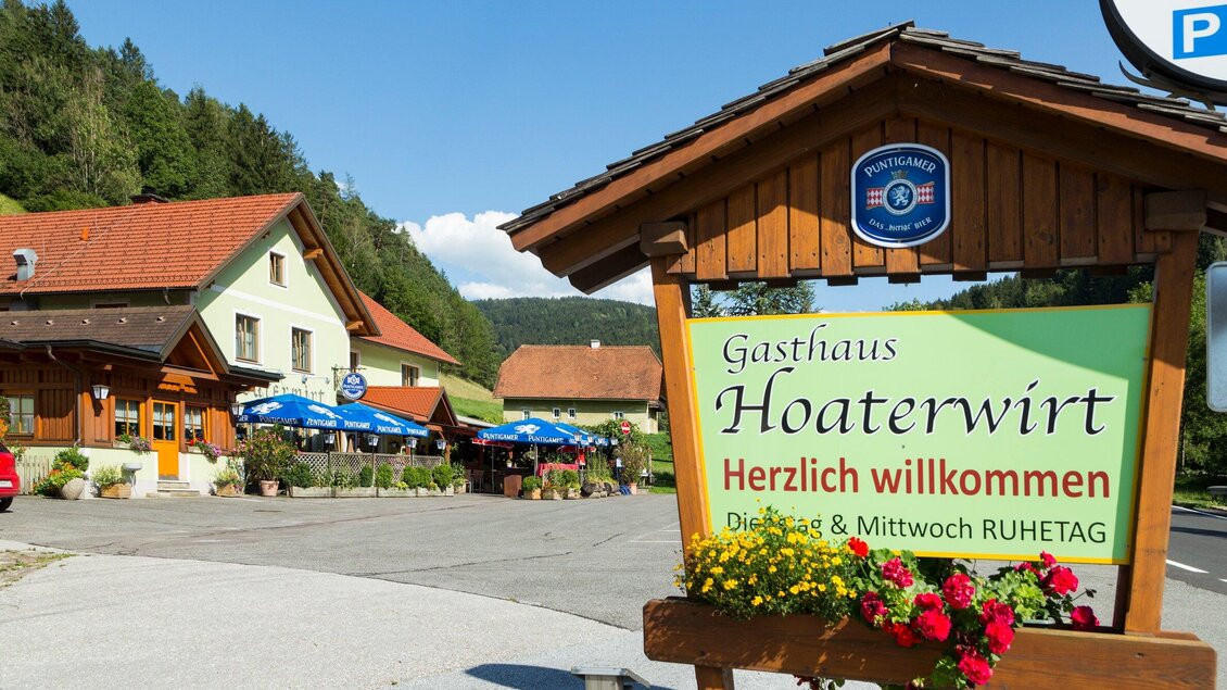 Ein einladendes Gasthaus mit einem Holzschild, das „Herzlich willkommen“ sagt. Im Hintergrund sieht man eine malerische Landschaft und Sonnenschirme, die zum Sitzen im Freien einladen. | © TV Region Graz - René Vidalli