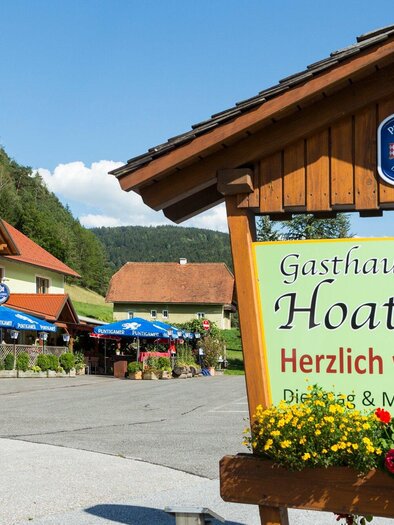 Ein einladendes Gasthaus mit einem Holzschild, das „Herzlich willkommen“ sagt. Im Hintergrund sieht man eine malerische Landschaft und Sonnenschirme, die zum Sitzen im Freien einladen. | © TV Region Graz - René Vidalli