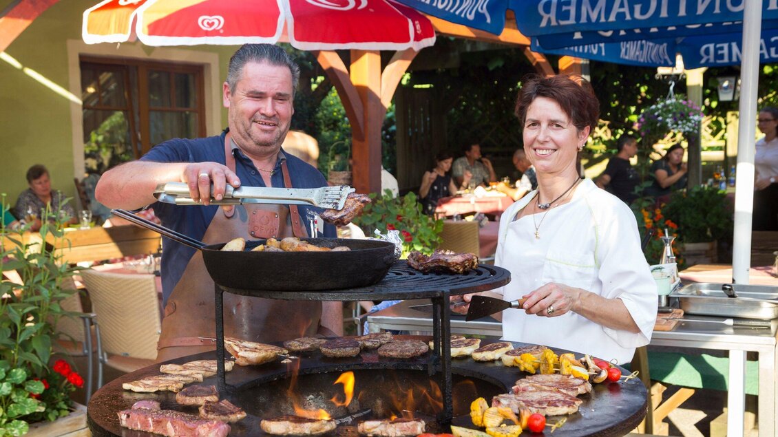 Ein Grillfest im Freien mit zwei lächelnden Personen. Sie bereiten Fleisch und Gemüse auf einem großen Grill zu. | © TV Region Graz - René Vidalli