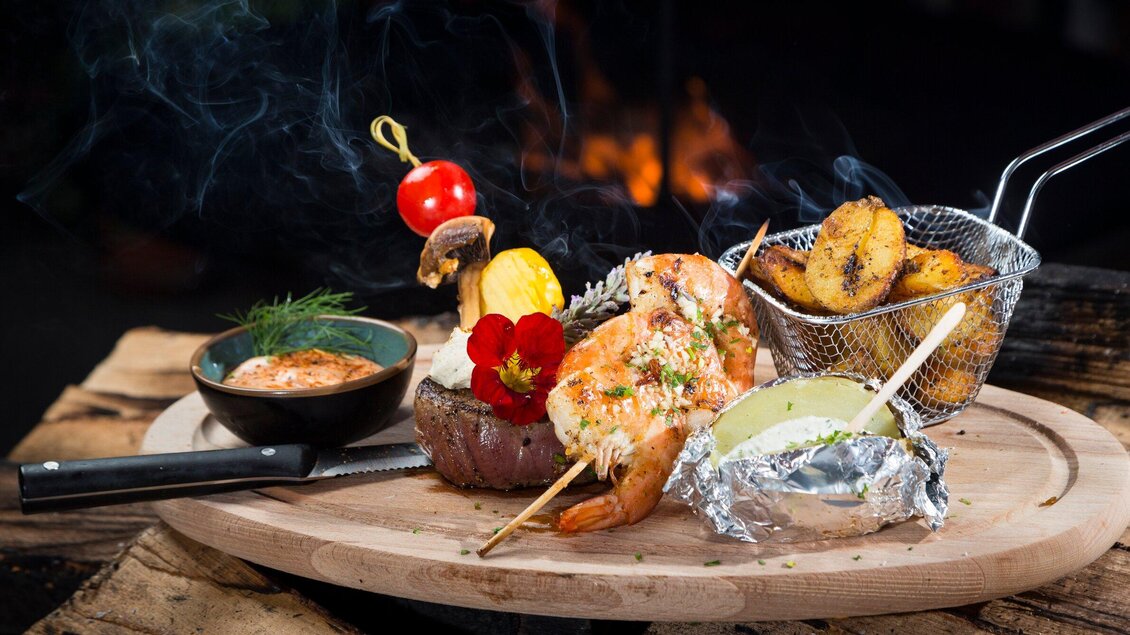 Ein köstliches Gericht auf einem Holzbrett, bestehend aus Garnelen, Fleisch, Beilagen und einer Sauce. Der Teller wird von frischen Kräutern und einer Tomate dekoriert. | © TV Region Graz - René Vidalli