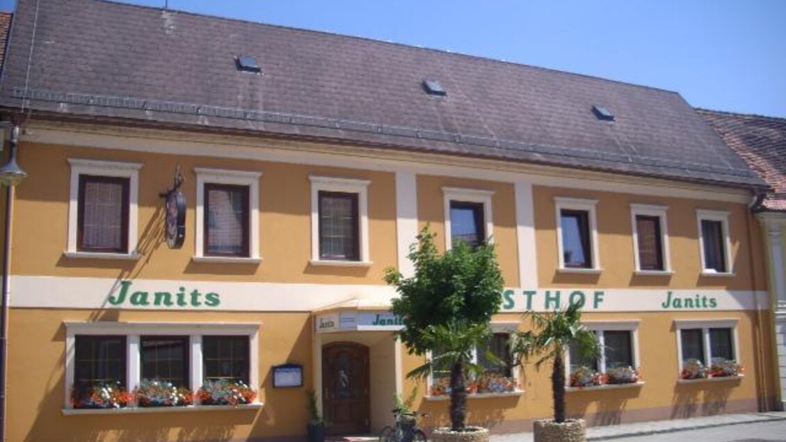Ein gelb gestrichenes Gebäude mit dem Schriftzug "Janits" und "Gasthof Janits". Vor dem Haus stehen Palmen und Blumen. | © Familie Janits