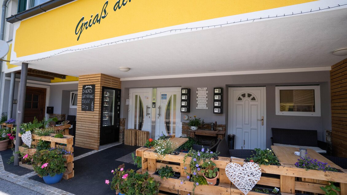 Ein einladendes Restaurant mit einem gelben Schild "Griaß di!" und blühenden Pflanzen. Der Außenbereich ist gemütlich gestaltet und bietet Platz zum Sitzen. | © Steirische Rucksackdörfer-KumpPhotography