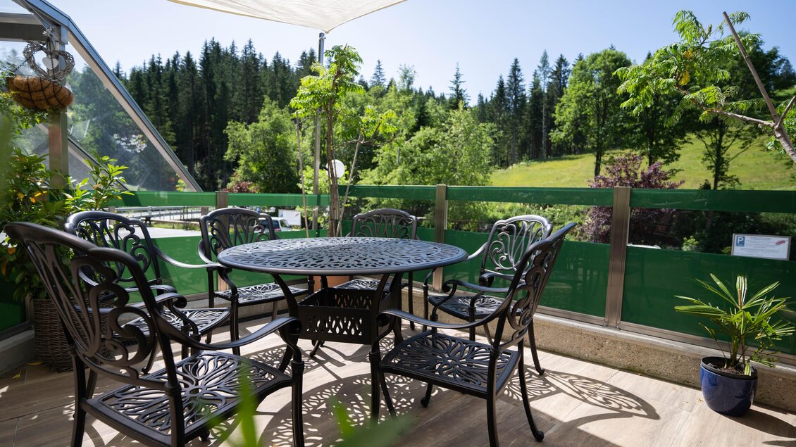 Ein gemütlicher Balkon mit einem runden Tisch und schwarzen Metallstühlen. Im Hintergrund sind grüne Bäume und eine sonnige Landschaft zu sehen. | © Kump