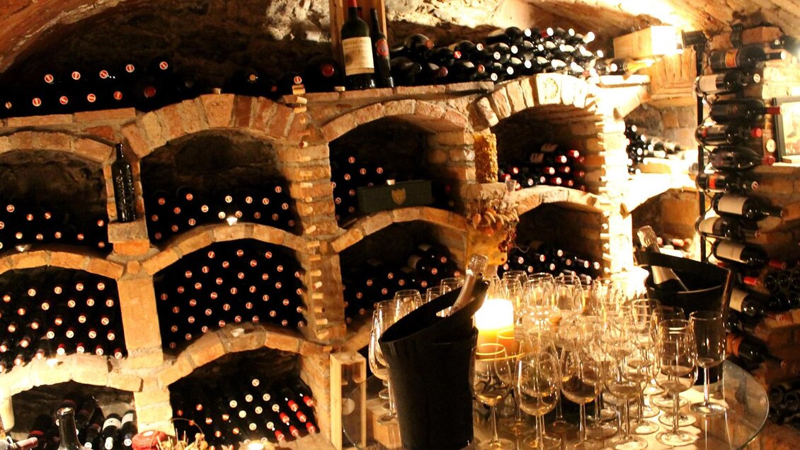 Ein Weinkeller mit Regalen voller Flaschen. In der Mitte steht ein Glastisch mit Sektkühler und Gläsern. | © Gasthof Knappenwirt