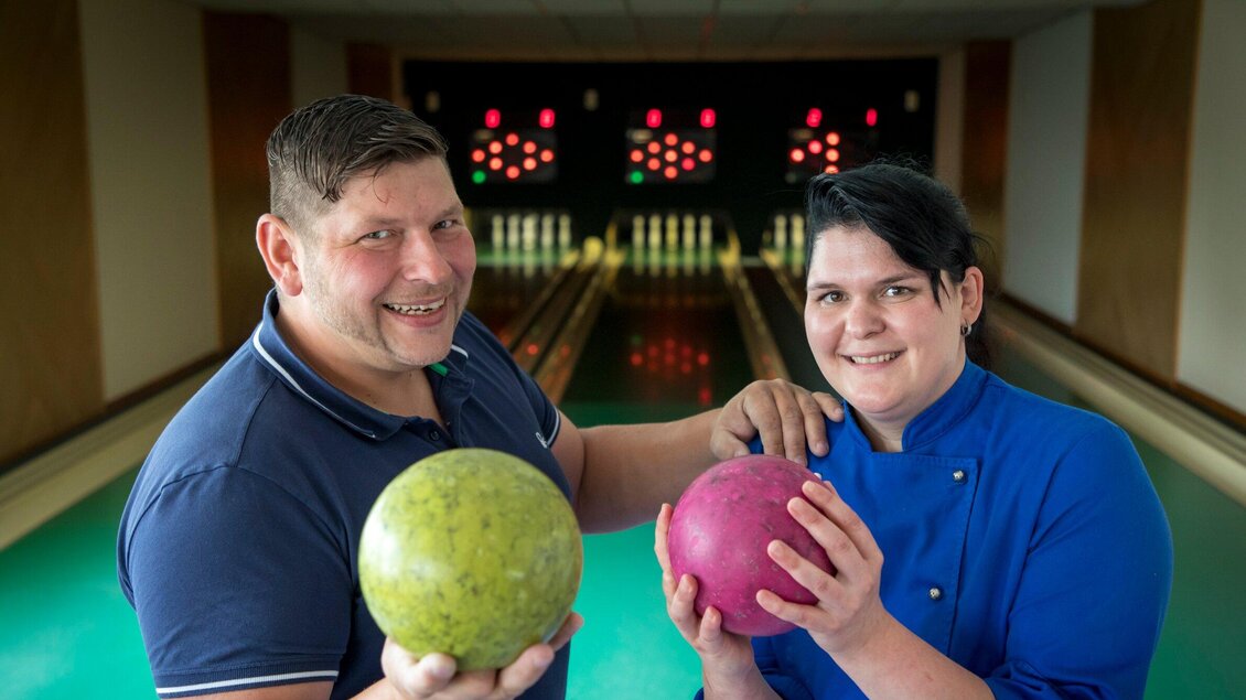 Zwei Personen stehen vor einer Bowlingbahn und halten jeweils einen Bowlingball in der Hand. Im Hintergrund sind die bunten Lichter der Bowlingbahn zu sehen. | © TV Region Graz - Lunghammer