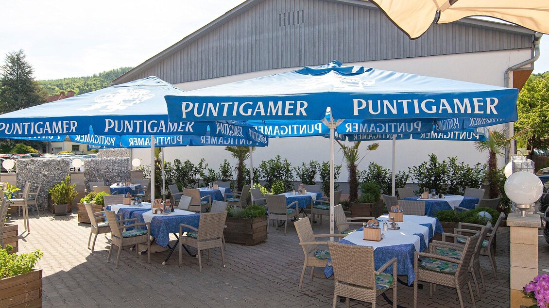 Eine gemütliche Terrasse mit blauen Tischdecken und Sonnenschirmen. Im Hintergrund sind Pflanzen und ein Gebäude sichtbar. | © TV Region Graz - Lunghammer