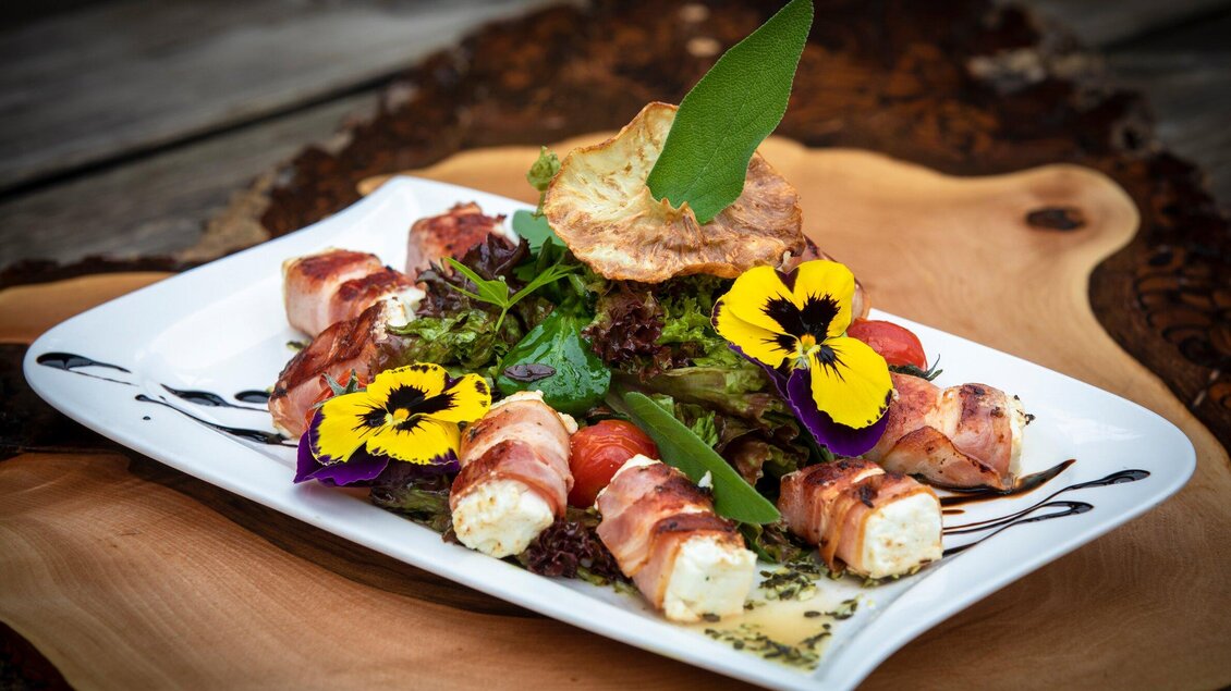 Ein ansprechend angerichteter Teller mit gefülltem Gemüse, umwickelt in Speck. Frische Salatblätter und essbare Blüten verleihen dem Gericht eine besondere Note. | © TV Region Graz - Lunghammer