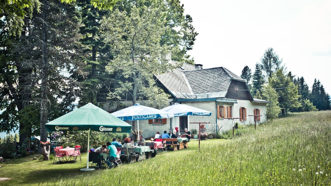 Ein rustikales Haus in einer grünen Wiese, umgeben von Bäumen. Mehrere Menschen sitzen unter Zelten und genießen die Natur. | © Lupi Spuma