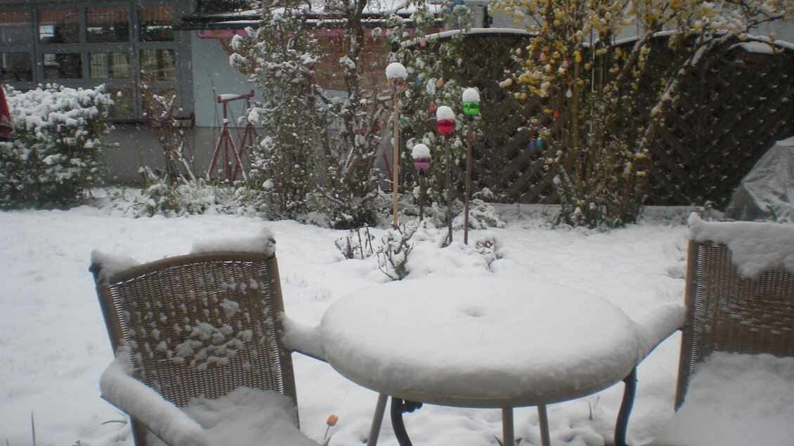 Ein verschneiter Garten mit einem Tisch und zwei Stühlen. Die Schneedecke bedeckt die Möbel und die Pflanzen im Hintergrund.