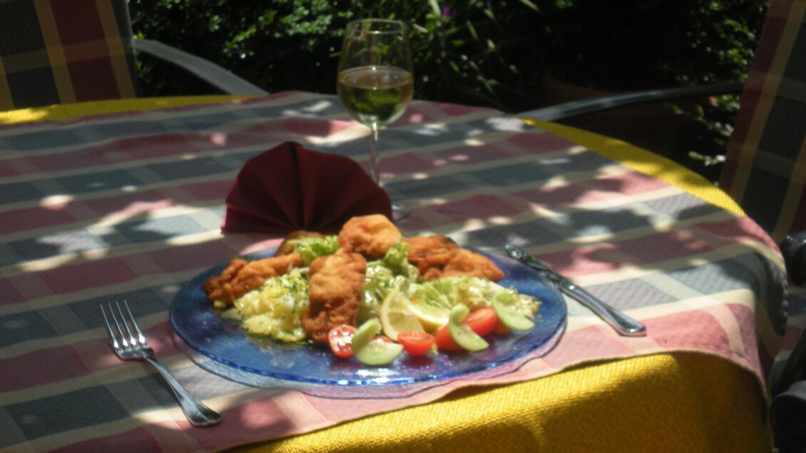 Ein gedeckter Tisch im Freien mit einem bunten Tischdecken. Auf dem Teller liegt eine Portion gebratenes Fleisch, Gemüse und ein Glas Wein.