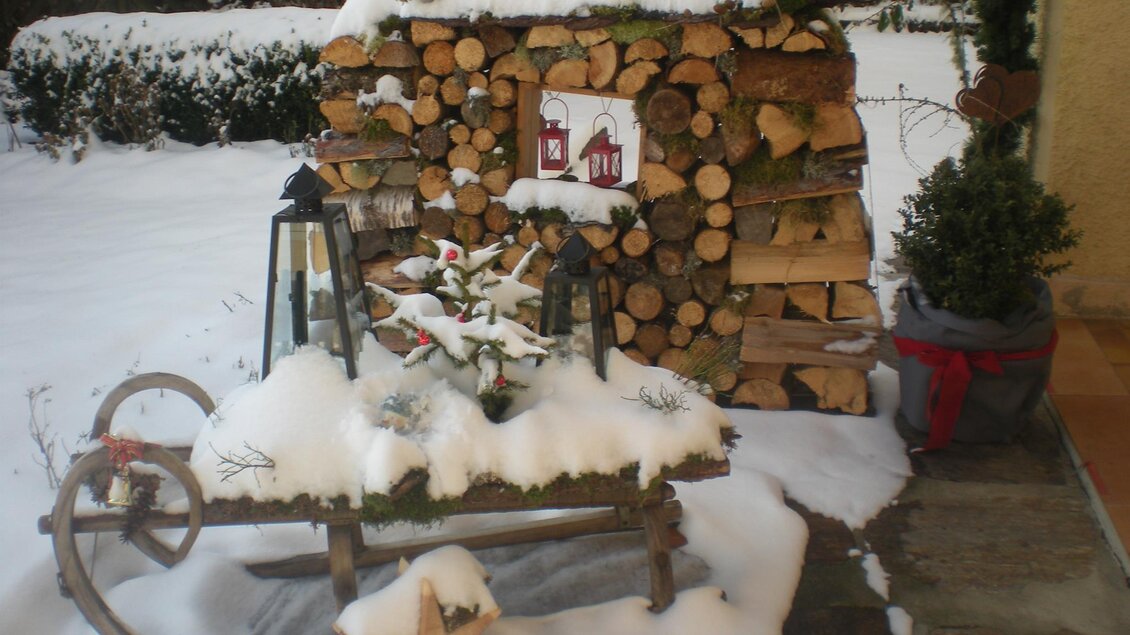 Ein gemütlicher Außenbereich im Schnee mit einem Holzstapel und einer dekorativen Bank. Auf der Bank sind Pflanzen und besinnliche Weihnachtsdekorationen.