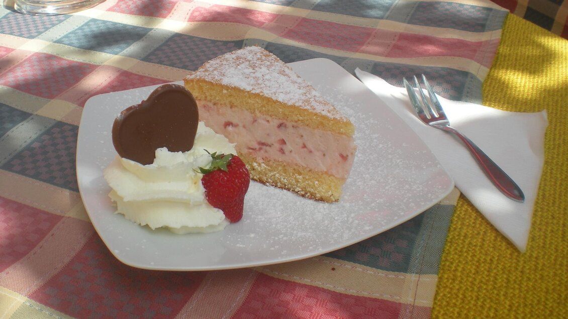 Ein Stück Erdbeertorte auf einem weißen Teller, dekoriert mit Schlagsahne und einer Erdbeere. Daneben liegt eine kleine, herzförmige Schokoladendekoration.