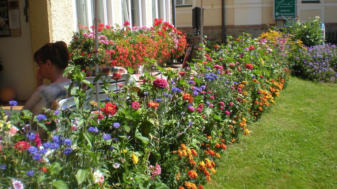 Ein bunten Blumengarten mit vielen verschiedenen Blumen in leuchtenden Farben. Im Hintergrund ist eine Terrasse mit Sonnenschirmen zu sehen.