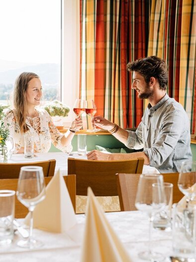 Ein Paar sitzt in einem gemütlichen Restaurant und stößt mit Gläsern an. Die Tische sind elegant gedeckt und es gibt Sonnenblumen auf dem Tisch. | © Martinhof