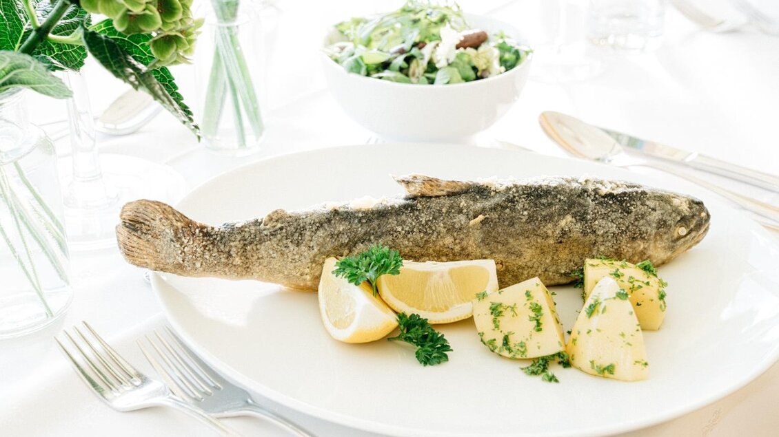 Ein frisch zubereiteter Fisch auf einem Teller, garniert mit Zitronenscheiben und Petersilie. Daneben liegen Kartoffeln und eine Schüssel Salat. | © Martinhof