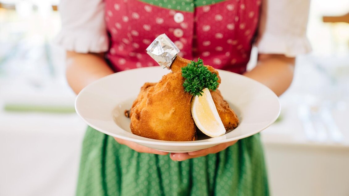 Eine Person trägt einen Teller mit einem großen, panierten Stück Fleisch, garniert mit einer Zitronenscheibe und Petersilie. Die Person ist in traditioneller bayerischer Tracht gekleidet. | © Martinhof