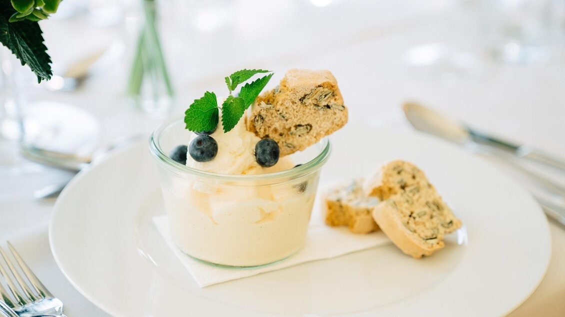 Ein köstliches Dessert in einem Glas mit Sahne, Minzblättern und Heidelbeeren. Daneben liegt ein Stück Kekse auf einem weißen Teller. | © Martinhof