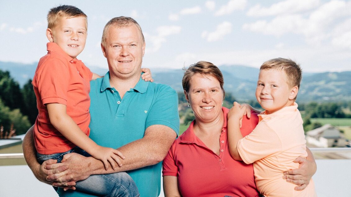 Eine Familie posiert im Freien, umgeben von Bergen und einer klaren Himmel. Die Eltern halten jeweils ein Kind in den Armen und lächeln glücklich. | © Martinhof