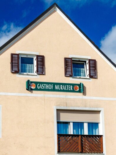 Ein traditionelles Gasthaus mit der Aufschrift "Gasthof Muralter". Die Fassade ist hell und freundlich, umgeben von einem blauen Himmel. | © TV Region Graz-Harry Schiffer