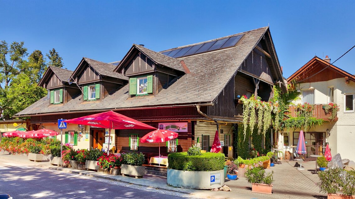 Ein charmantes Holzhaus mit grünen Fensterläden und roten Schirmen im Vordergrund. Die Umgebung ist grün und einladend, ideal für einen gemütlichen Aufenthalt. | © Gasthof Kranerwirt
