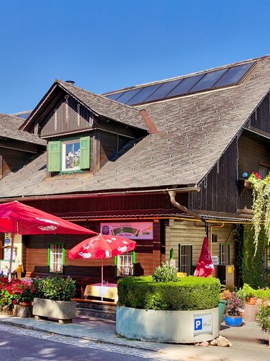 A charming wooden house with green shutters and red umbrellas in the foreground. The surroundings are green and inviting, perfect for a cozy stay. | © Gasthof Kranerwirt
