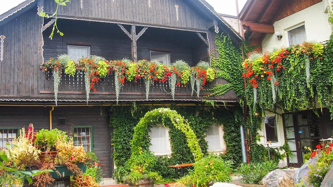 Ein charmantes Haus mit Holzverkleidung und üppigen Pflanzen. Bunte Blumen hängen in Kästen und verdecken teilweise die Fassade. | © Gasthof Niggas "Kranerwirt"