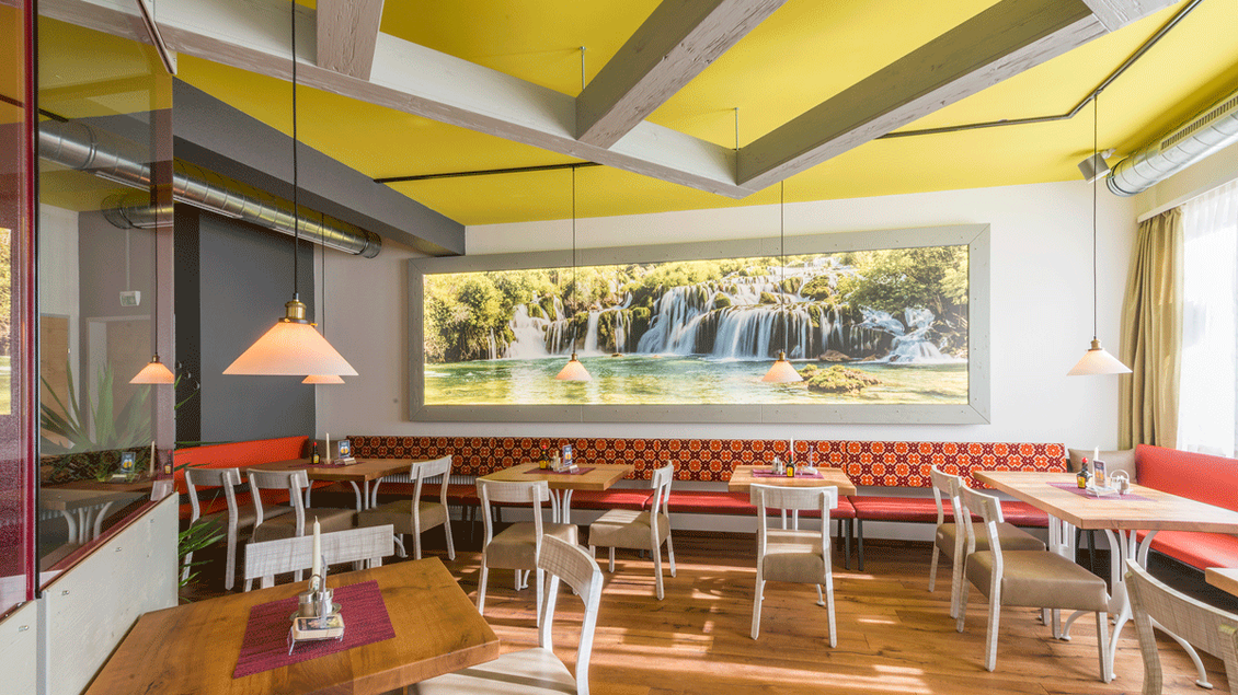 Ein modernes Restaurant mit hellen Wänden und Holzfußboden. Große Fenster lassen viel Licht herein, und ein Wandbild zeigt einen ruhigen Wasserfall. | © TV Hochsteiermark