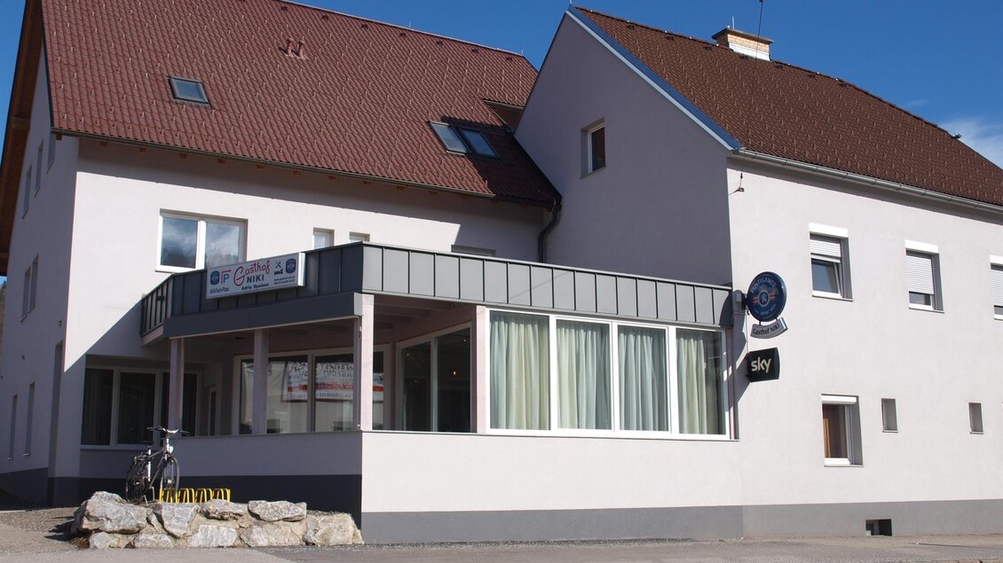 Ein großes, helles Gebäude mit einem roten Satteldach und einem modernen Anbau. Vor dem Gebäude stehen einige Steine und ein Fahrrad ist sichtbar. | © TV Hochsteiermark