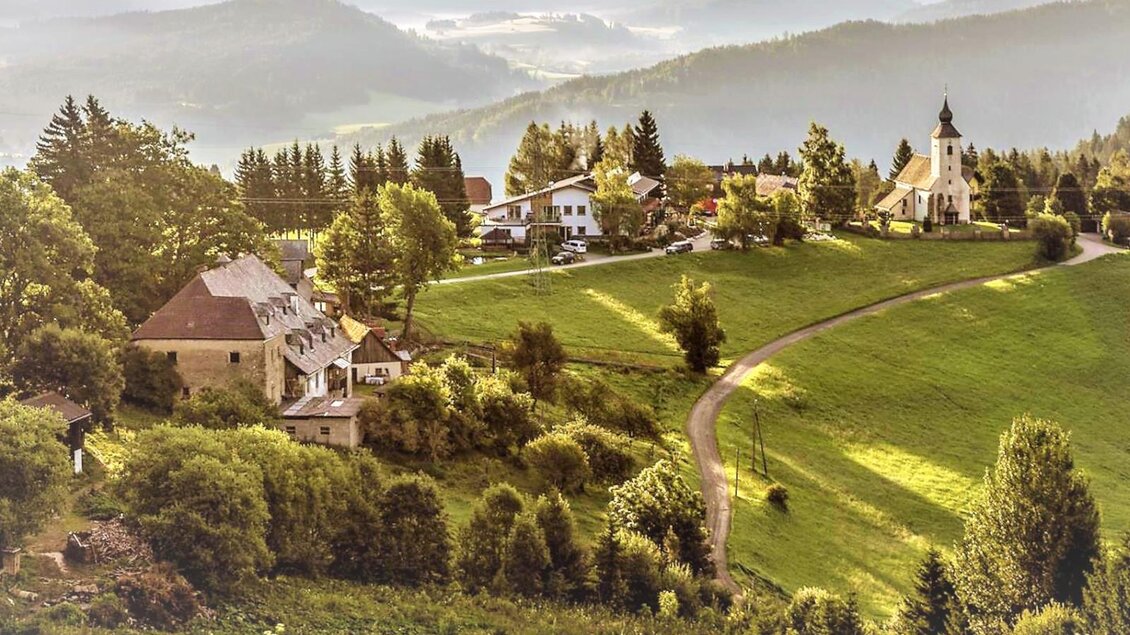 Eine ruhige, ländliche Schau mit grünen Hügeln und verstreuten Häusern. Im Hintergrund ist eine kleine Kirche und sanfte Berge zu sehen. | © Gasthof Pöllauerhof