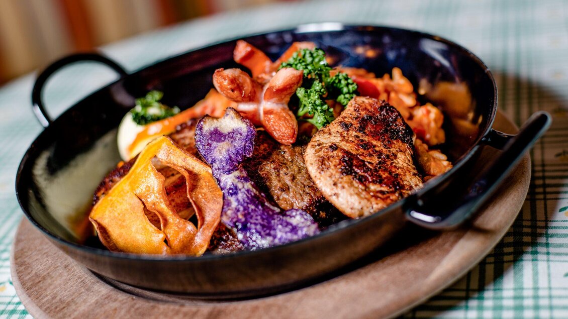 Ein kunstvoll angerichteter Teller mit gegrilltem Fleisch, buntem Gemüse und knusprigen Chips. Das Gericht ist in einer schwarzen Schüssel auf einem Holzbrett präsentiert. | © TV Region Graz - Mias Photoart