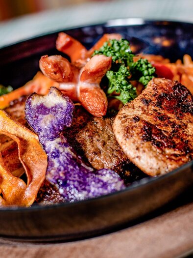 Ein kunstvoll angerichteter Teller mit gegrilltem Fleisch, buntem Gemüse und knusprigen Chips. Das Gericht ist in einer schwarzen Schüssel auf einem Holzbrett präsentiert. | © TV Region Graz - Mias Photoart