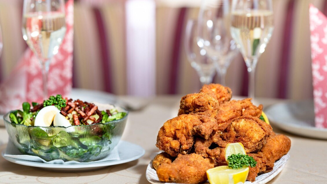 Ein Teller mit knusprigen Hähnchenstücken und eine Schüssel mit frischem Salat. Im Hintergrund sind Gläser mit Getränken zu sehen. | © TV Region Graz - Mias Photoart