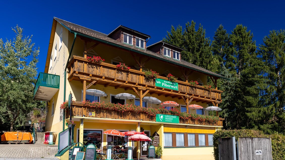 Ein charmantes Hotelgebäude mit einem Holzbalkon und Blumen. Vor dem Eingang stehen Tische mit Sonnenschirmen. | © Lipizzanerheimat | Harry Schiffer