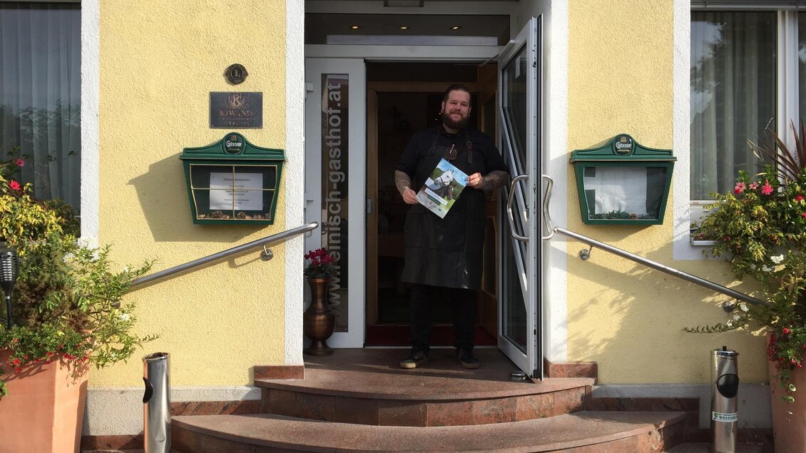 Ein Mann steht vor einem Restaurant mit gelber Fassade und begrüßt die Gäste. Er hält eine Speisekarte in der Hand und die Umgebung ist freundlich gestaltet mit Pflanzen. | © TV Lipizzanerheimat