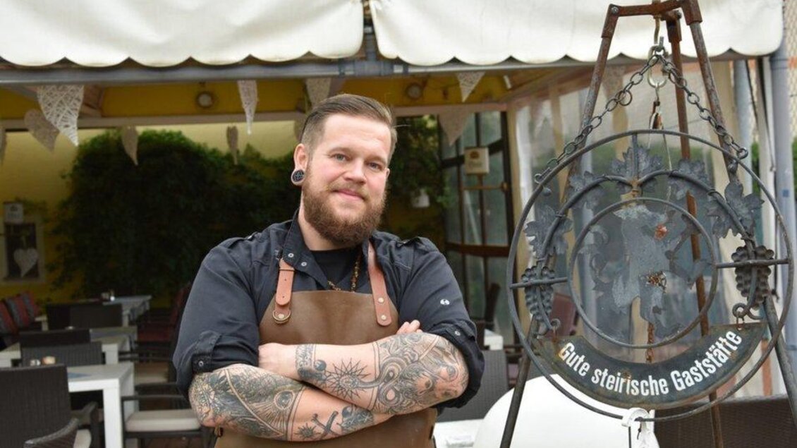 Ein junger Mann mit Bart und Tattoos steht in einem Restaurant mit gemütlicher Terrasse. Er trägt eine Schürze und lächelt freundlich in die Kamera. | © Fam. Reinisch
