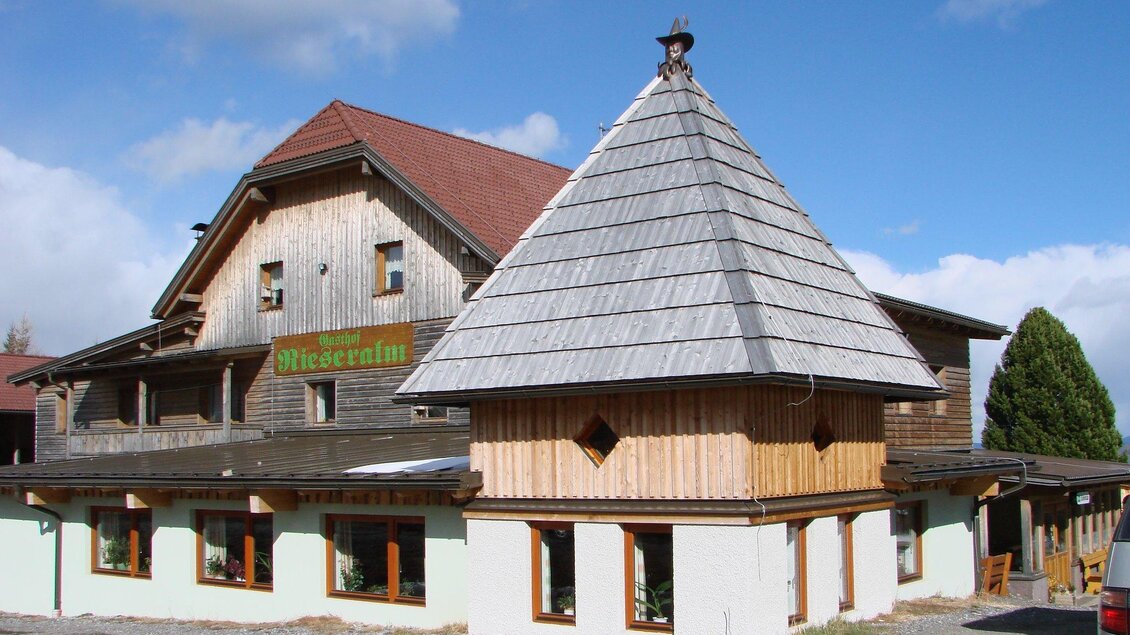 Ein traditionelles Holzgebäude mit einem charakteristischen Schindeldach. Das Gebäude hat große Fenster und ist von einer ruhigen Umgebung umgeben. | © Gasthof Rieseralm