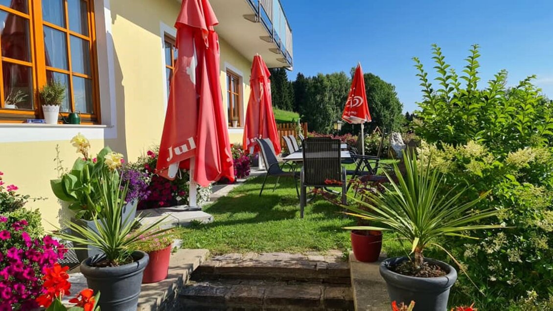 Ein schöner Gartenbereich mit bunten Blumen und grünen Pflanzen. Auf der Terrasse stehen rote Sonnenschirme und bequeme Gartenmöbel. | © Gasthof Roschitzhof