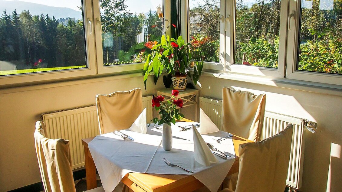 Ein gemütlicher Tisch mit weißen Tischdecken und Blumen in einer sonnigen Ecke. Durch die Fenster sieht man eine grüne Landschaft und Bäume. | © GH Safran Filatsch