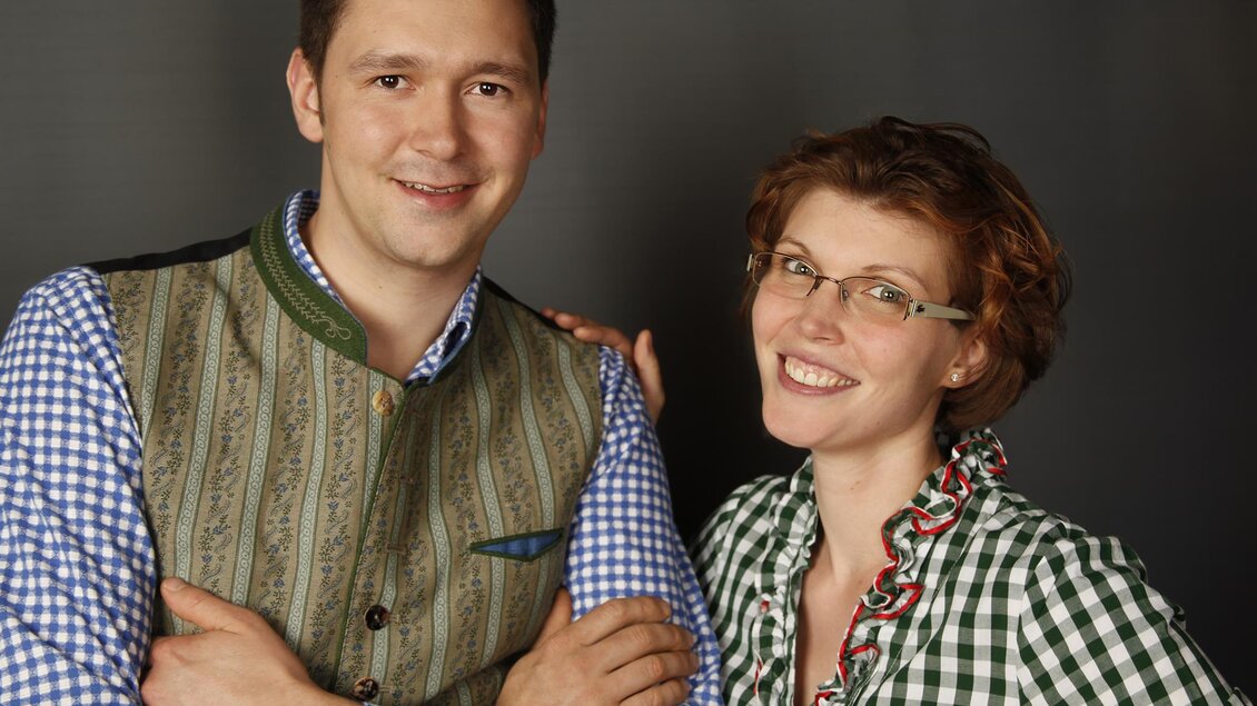 Ein Paar in traditioneller bayerischer Kleidung lächelt freundlich. Der Mann trägt eine Weste, während die Frau ein kariertes Hemd hat. | © GH Schäffer