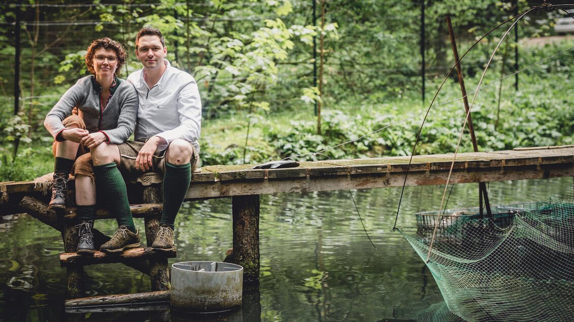 Ein Paar sitzt auf einem Holzsteg über einem ruhigen Gewässer. Im Hintergrund sind grüne Bäume und eine Fischernetz zu sehen. | © GH Schäffer_Neuberg (C) Seiser_Karner