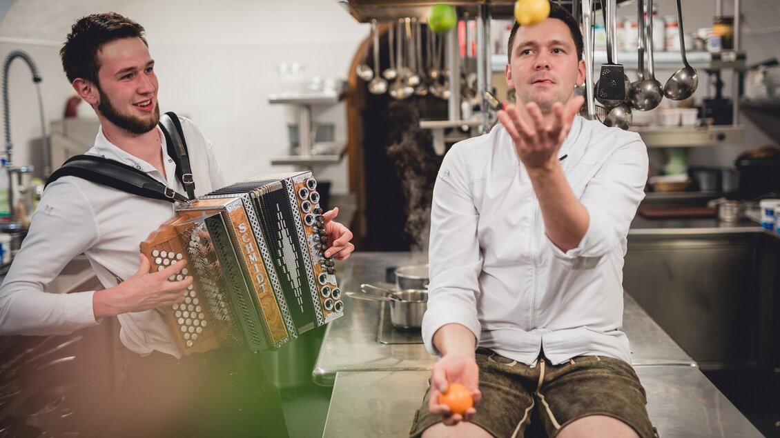 Zwei Männer in einer Küche, einer spielt Akkordeon und der andere jongliert mit Obst. Eine fröhliche Atmosphäre in einem gastronomischen Umfeld. | © GH Schäffer_Neuberg (C) Seiser_Karner