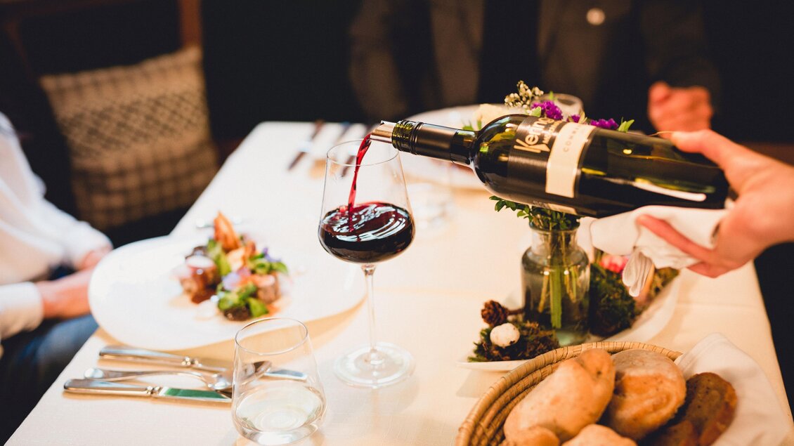 Ein elegantes Restaurant-Setting mit einem Glas Rotwein, das gerade eingeschenkt wird. Im Hintergrund sind ein Teller mit Salat und frisches Brot zu sehen. | © GH Schäffer_Neuberg (C) Seiser_Karner