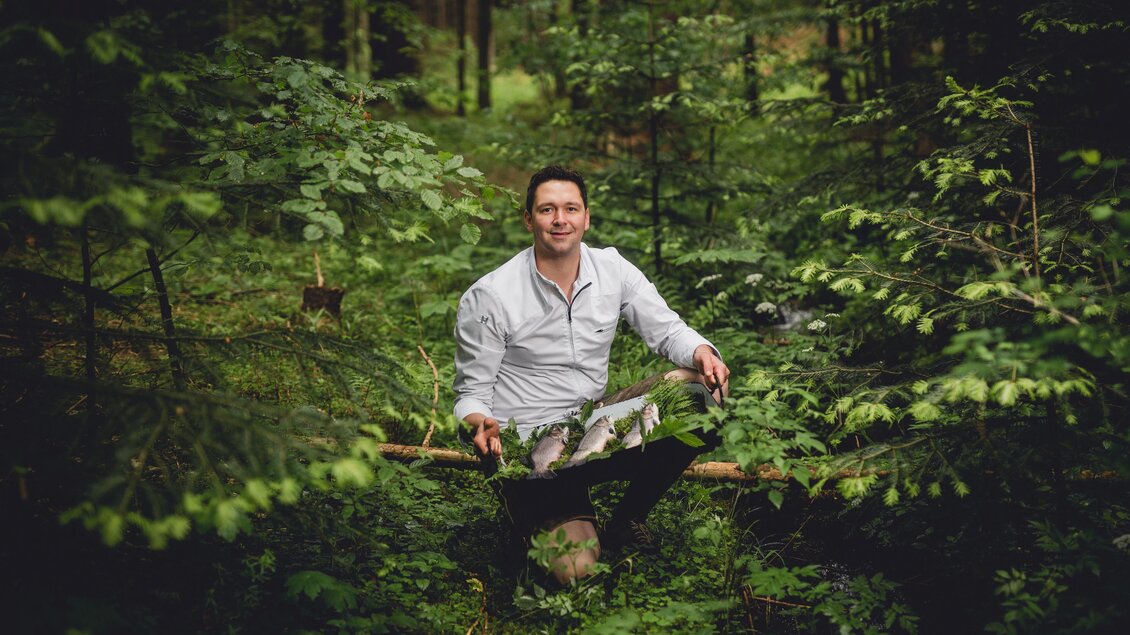 Ein Mann sitzt entspannt im Wald auf einem Baumstamm. Um ihn herum wachsen viele grüne Pflanzen und Bäume. | © GH Schäffer_Neuberg (C) Seiser_Karner