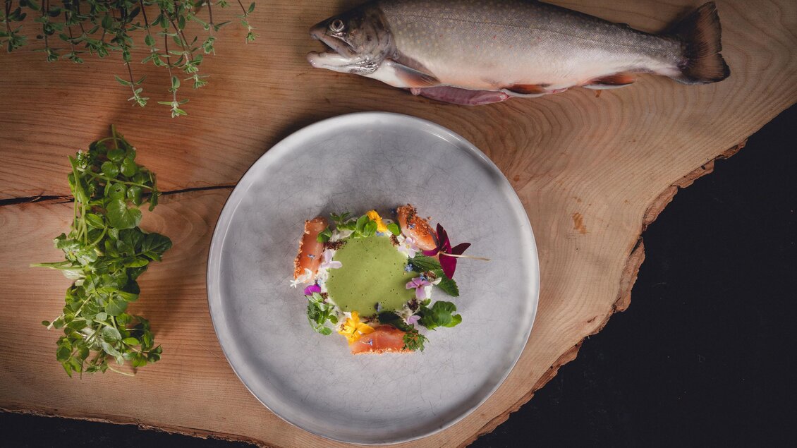 Ein elegantes Gericht auf einem Teller mit frischen Zutaten und essbaren Blumen. Im Hintergrund liegt ein ganzer Fisch auf einem Holzbrett. | © GH Schäffer_Neuberg (C) Seiser_Karner