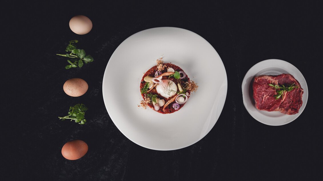Ein elegantes Gericht auf einem weißen Teller, garniert mit Meeresfrüchten und frischem Gemüse. Neben dem Teller liegen einige Eier und eine Schale mit rohem Fleisch. | © GH Schäffer_Neuberg (C) Seiser_Karner