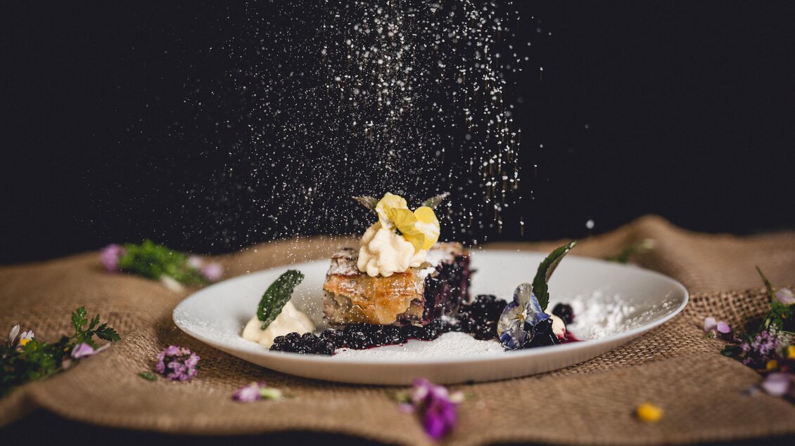 Ein köstliches Dessert auf einem Teller, dekoriert mit Sahne und Beeren. Feiner Puderzucker wird darüber gestreut und umgeben von bunten Blüten. | © GH Schäffer_Neuberg (C) Seiser_Karner